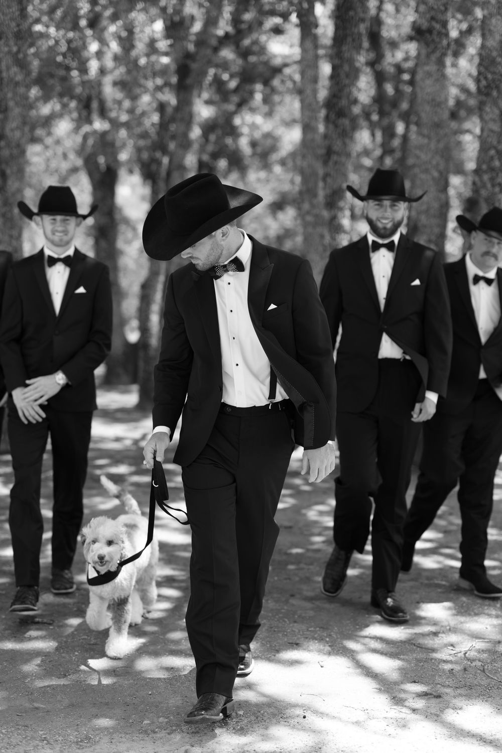 Black and white photo of groomsmen in western attire walking with a small white dog in the woods.