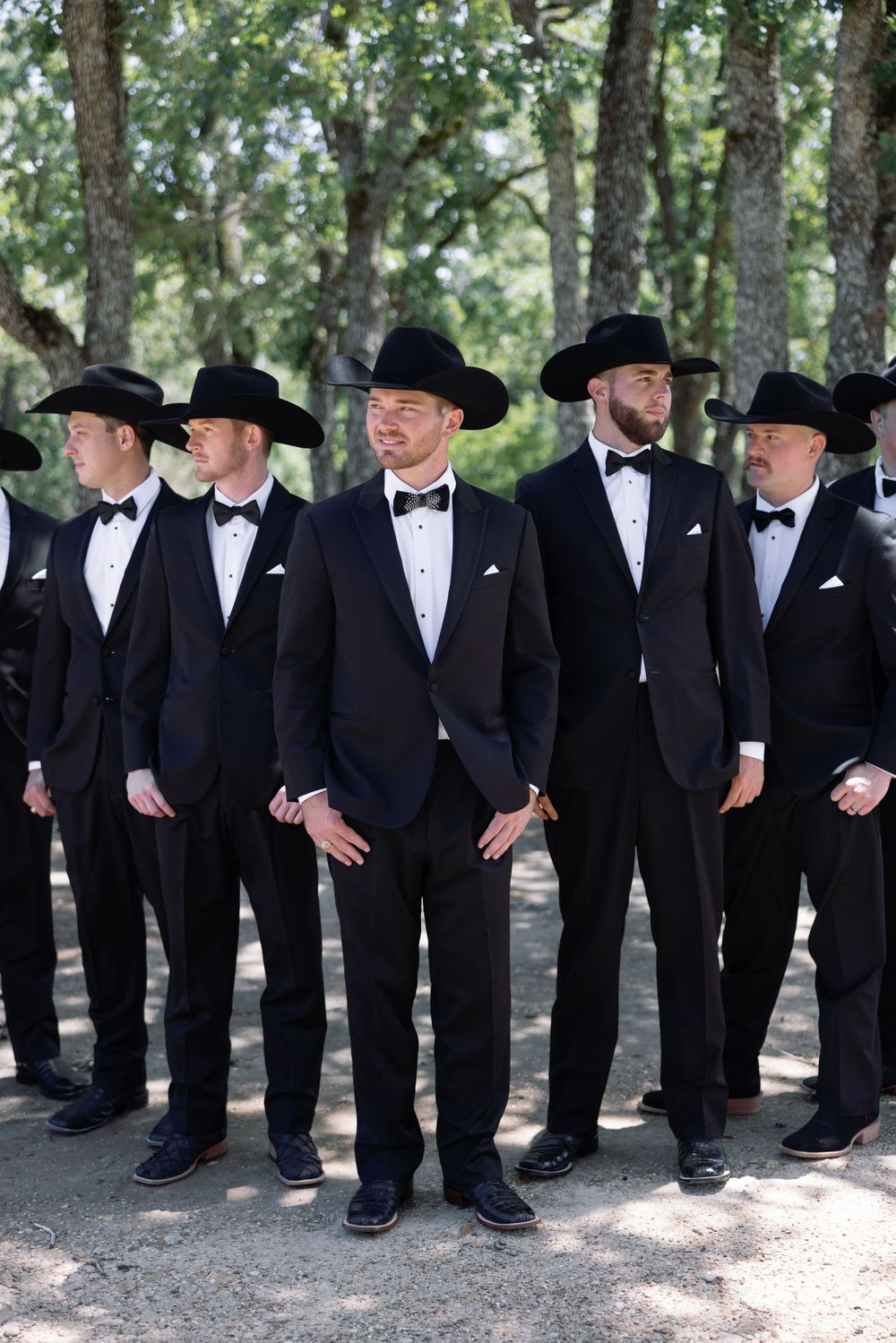 Groomsmen lineup wearing matching black tuxedos and cowboy hats at outdoor wedding.