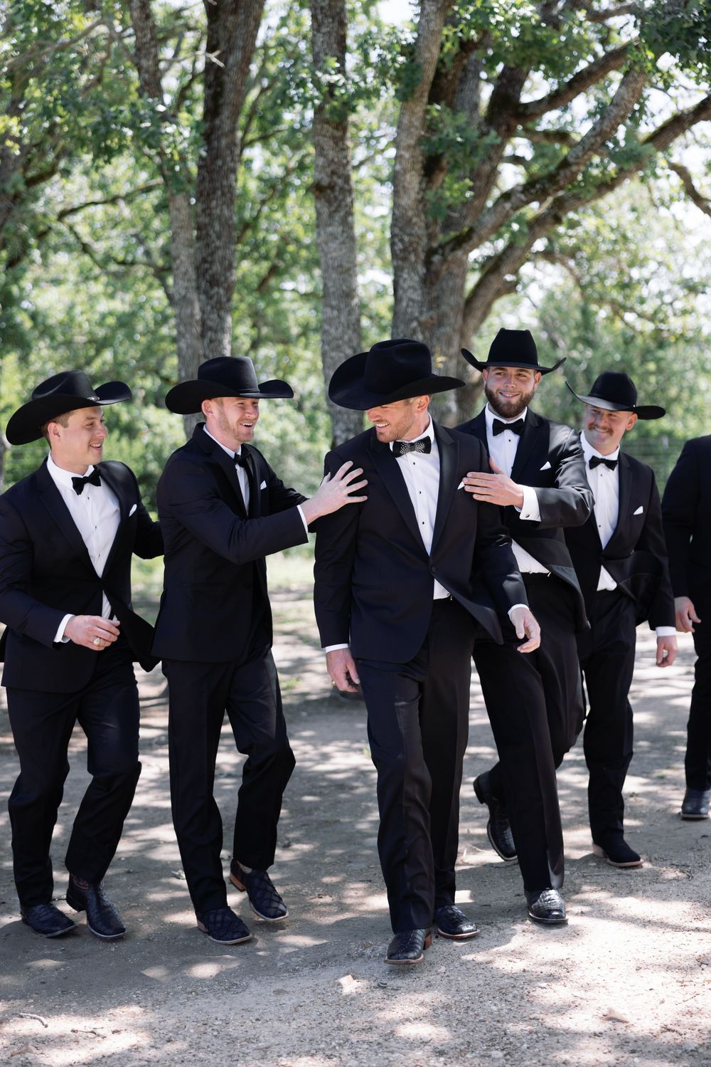 Groomsmen in black tuxedos and cowboy hats sharing a lighthearted moment outdoors under tree shade.