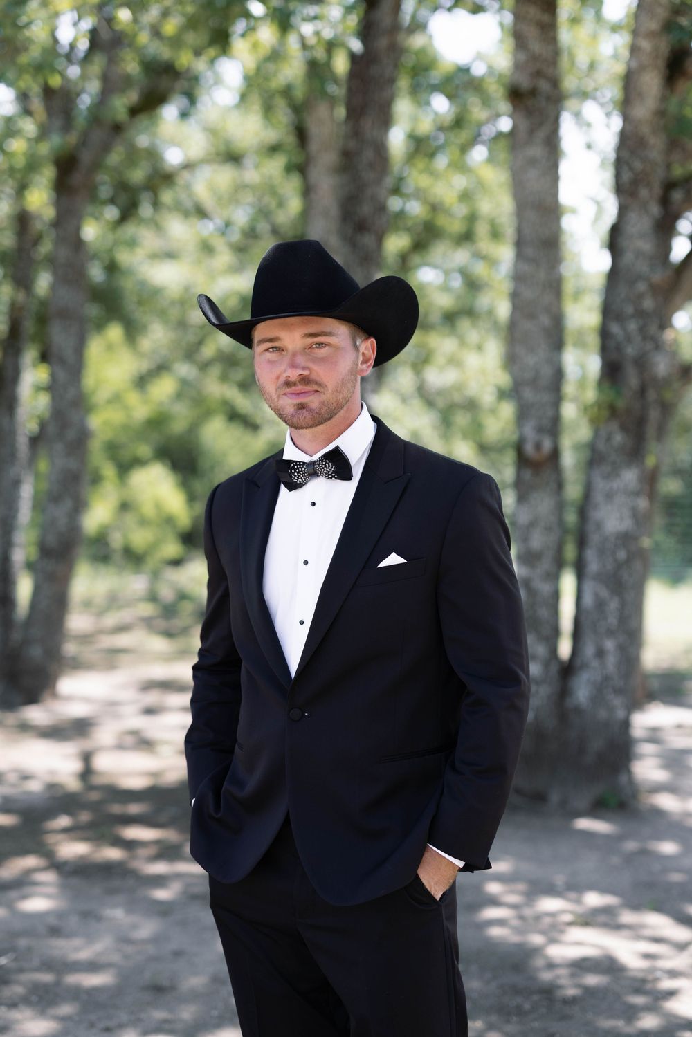 Groomsman in black tuxedo and cowboy hat poses for formal portrait against trees in outdoor setting.
