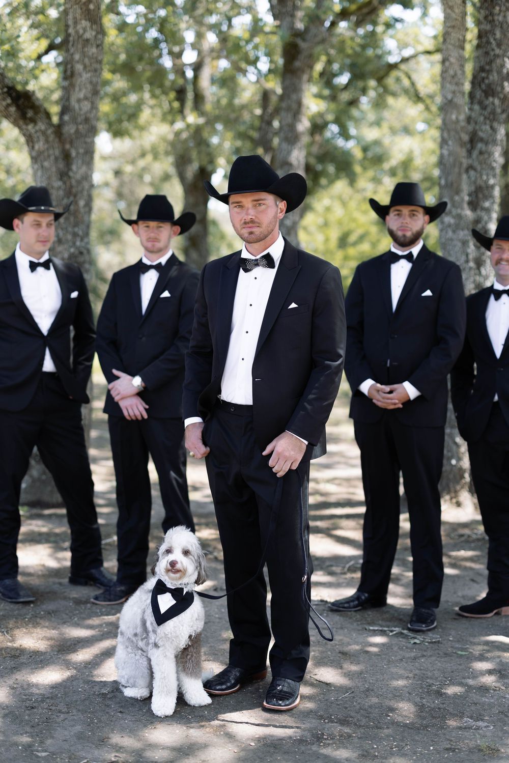 Groomsmen in black tuxedos and cowboy hats stand in a wooded area with a small white dog wearing a bowtie.