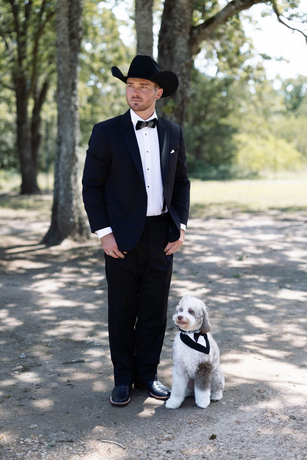 A man in a black tuxedo and cowboy hat stands outdoors with a small white dog wearing a bow tie.