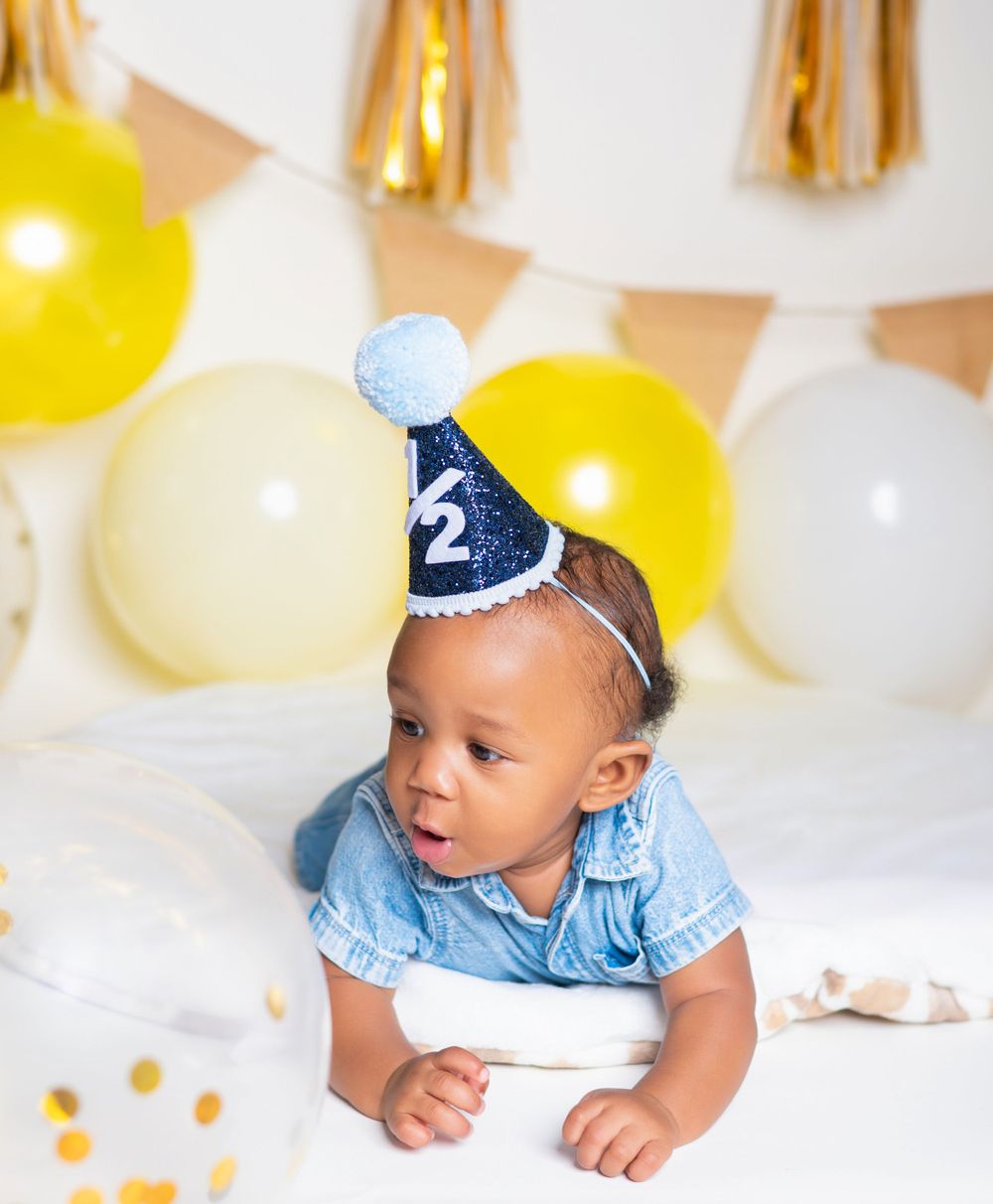 Baby wearing blue party hat and romper surrounded by yellow and white balloons.