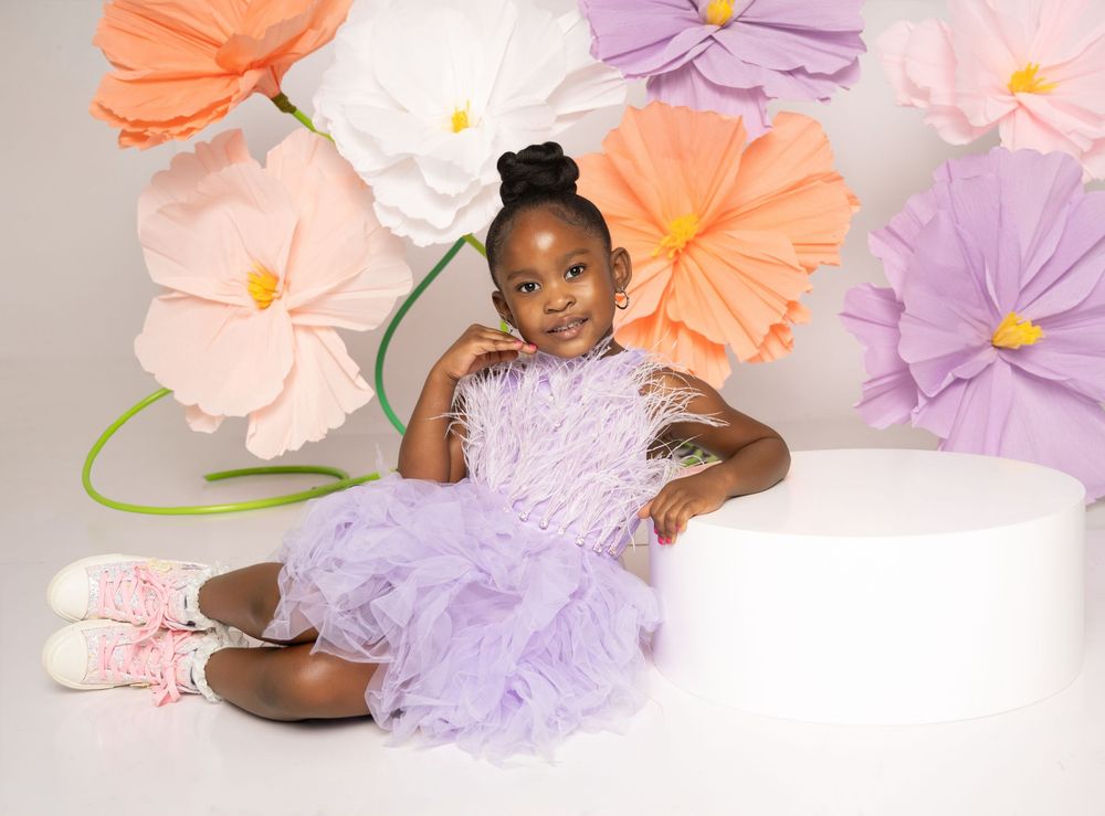 Young girl in lavender tutu dress poses in front of colorful paper flowers backdrop for a professional photo shoot.