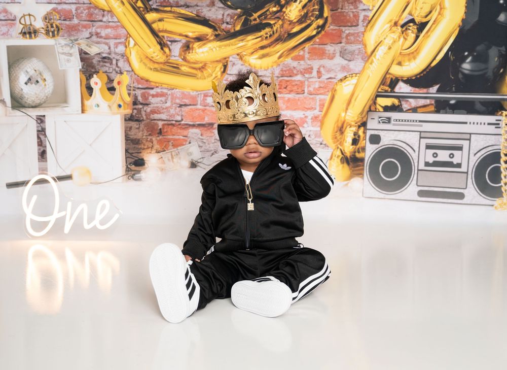 Adorable baby in black tracksuit and gold crown sunglasses sits for a first birthday photo shoot with boombox backdrop.