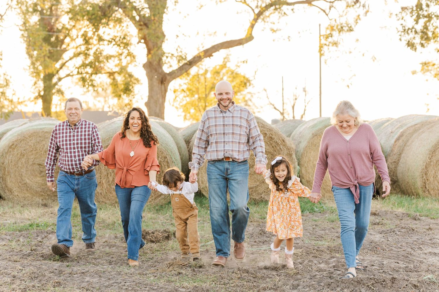 A multi-generational group walks together on a dirt path with round hay bales visible in the autumn field behind them.