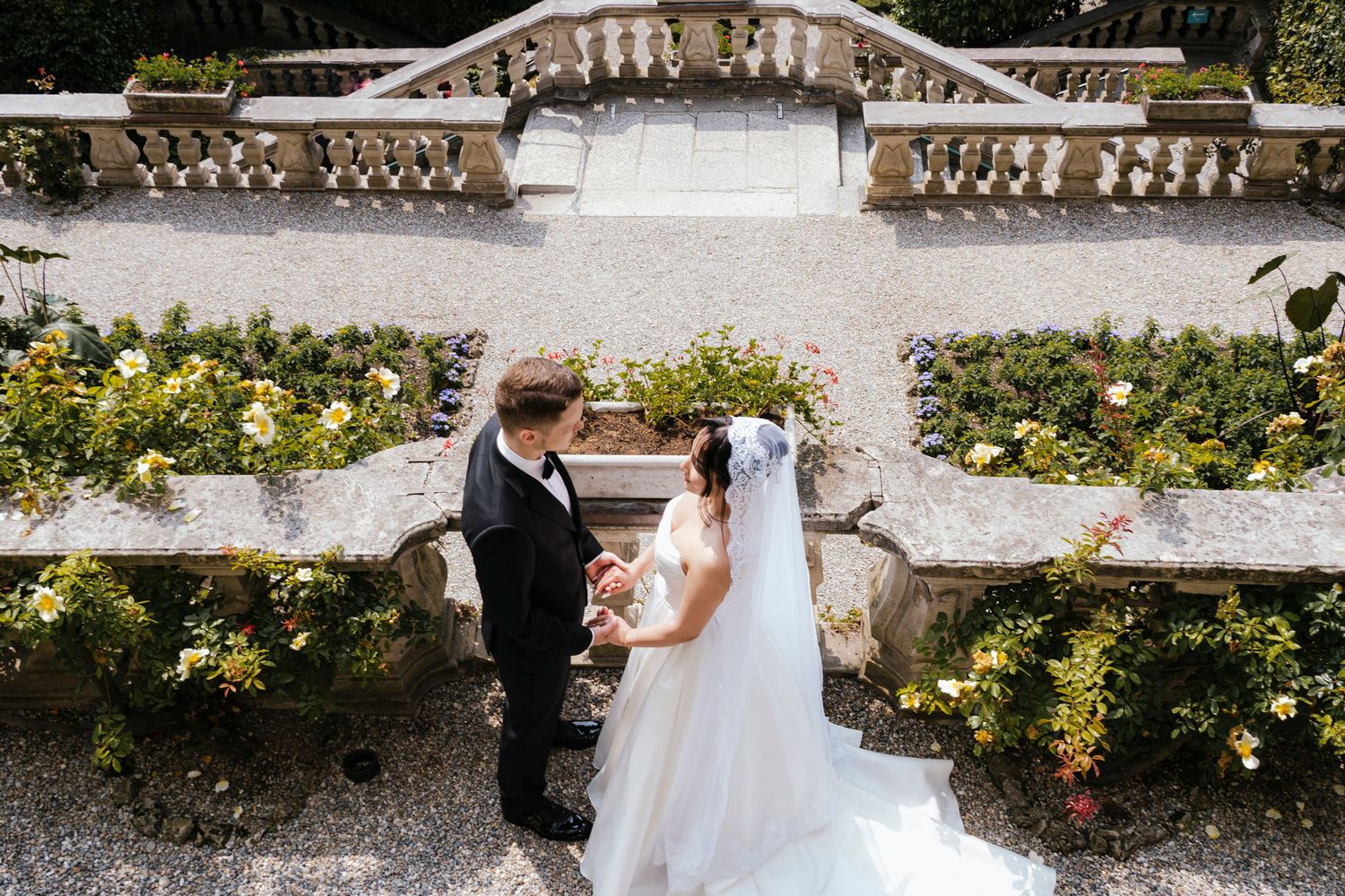 Couple in wedding attire walking hand in hand through a scenic garden with stone steps and vibrant foliage.