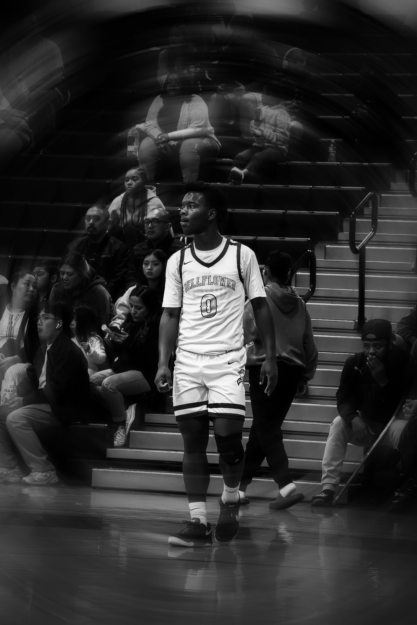 Basketball player in white jersey stands on gymnasium bleachers during black and white photo.