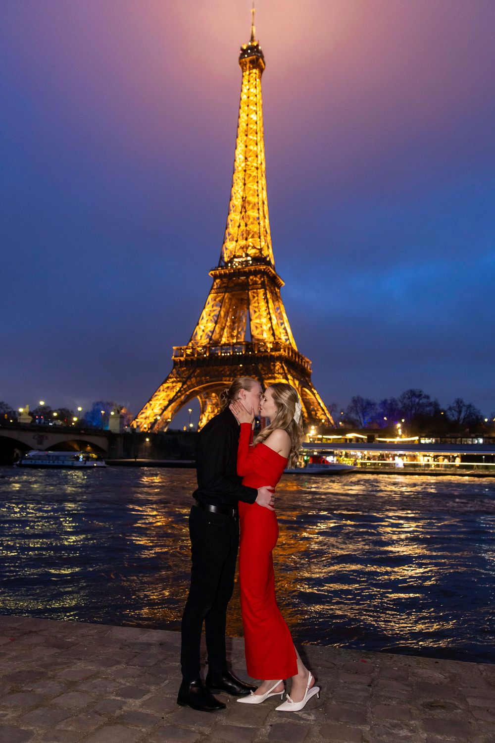 A couple embraces in front of the illuminated Eiffel Tower at night with the Seine River reflecting golden lights.