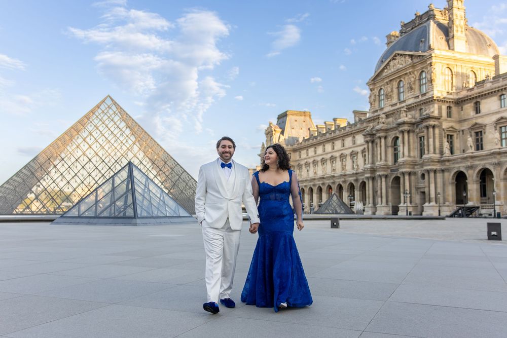 A couple in formal attire poses at the Louvre Museum plaza with its iconic glass pyramid in the background.