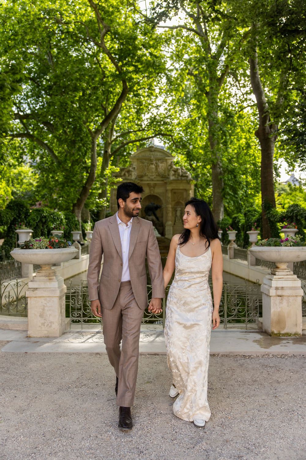 Couple walking hand in hand through a garden pathway with stone balustrades and lush green trees in elegant wedding attire.