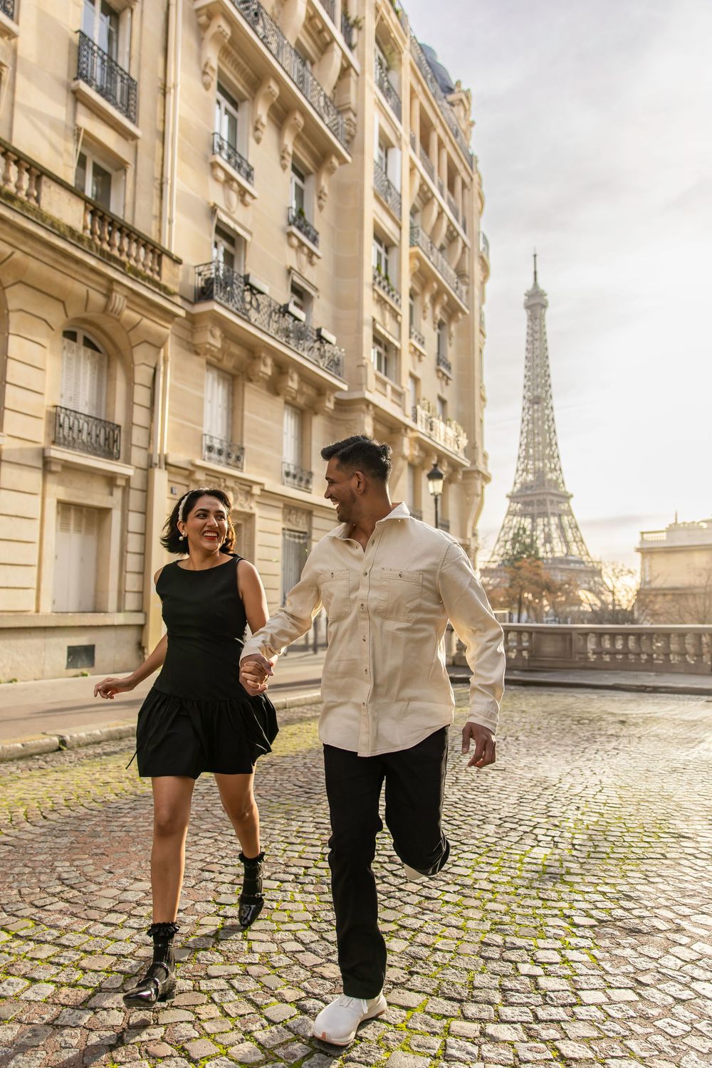 A couple runs joyfully on a cobblestone street in Paris with the Eiffel Tower visible in the background.
