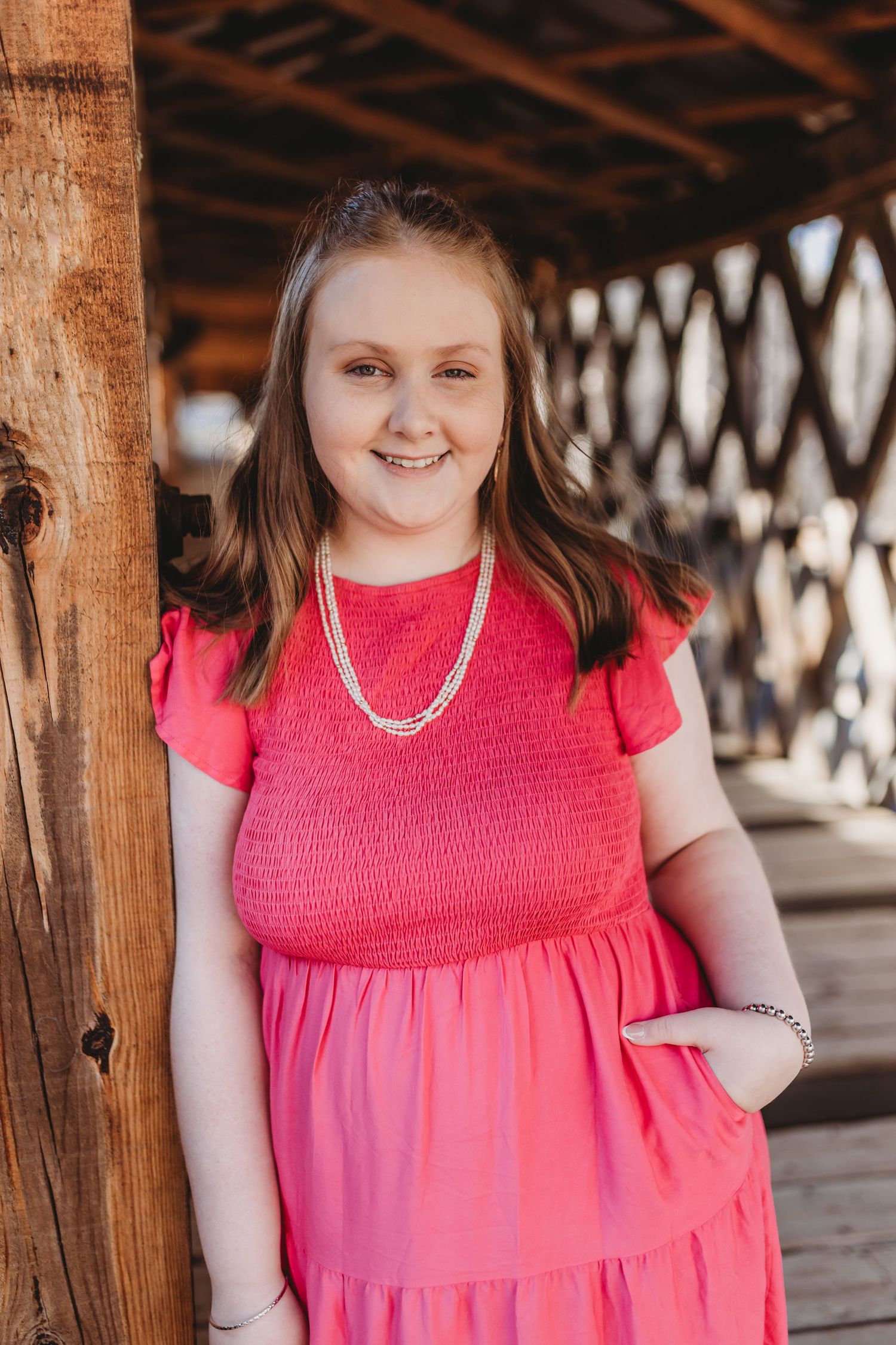 A person wearing a bright pink dress poses under a wooden covered bridge structure.