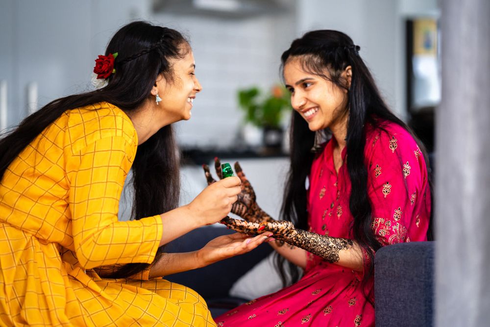 Two women in traditional Indian attire share a joyful moment during a mehndi ceremony with henna application.