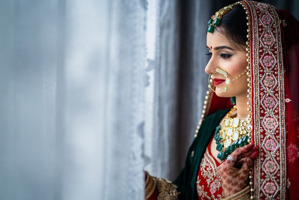 A bride in traditional Indian wedding attire poses elegantly in a red and gold lehenga with intricate embroidery and jewelry.
