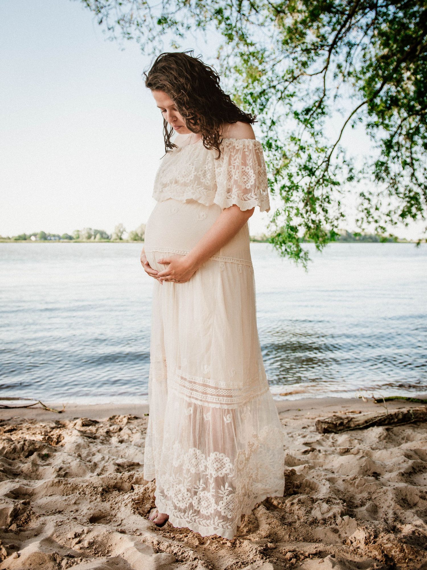 Pregnant woman in white lace off-shoulder maxi dress standing on sandy beach by water, cradling her baby bump.