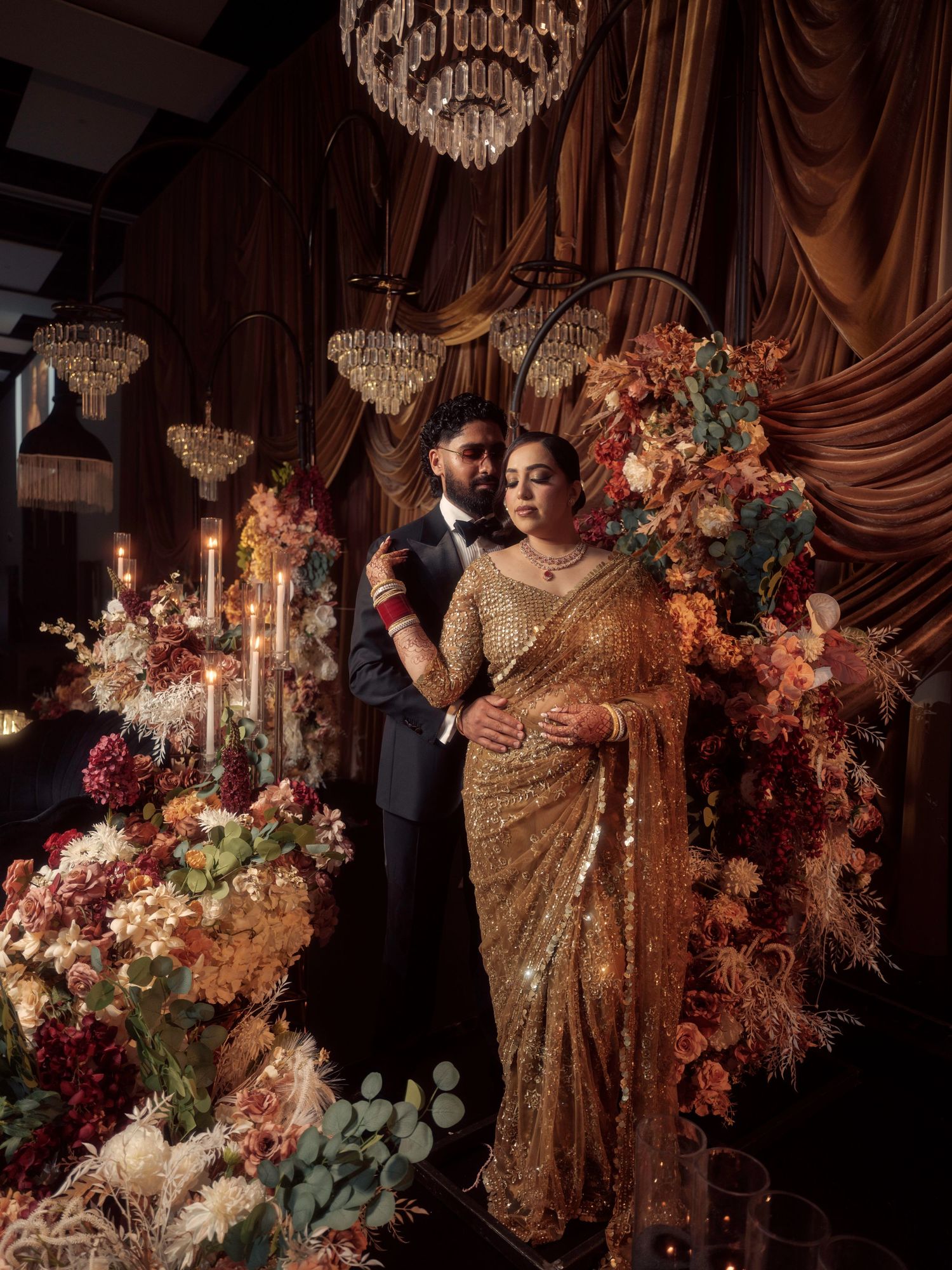 Elegant wedding couple poses in front of ornate crystal chandelier and lush floral arrangements in romantic setting.