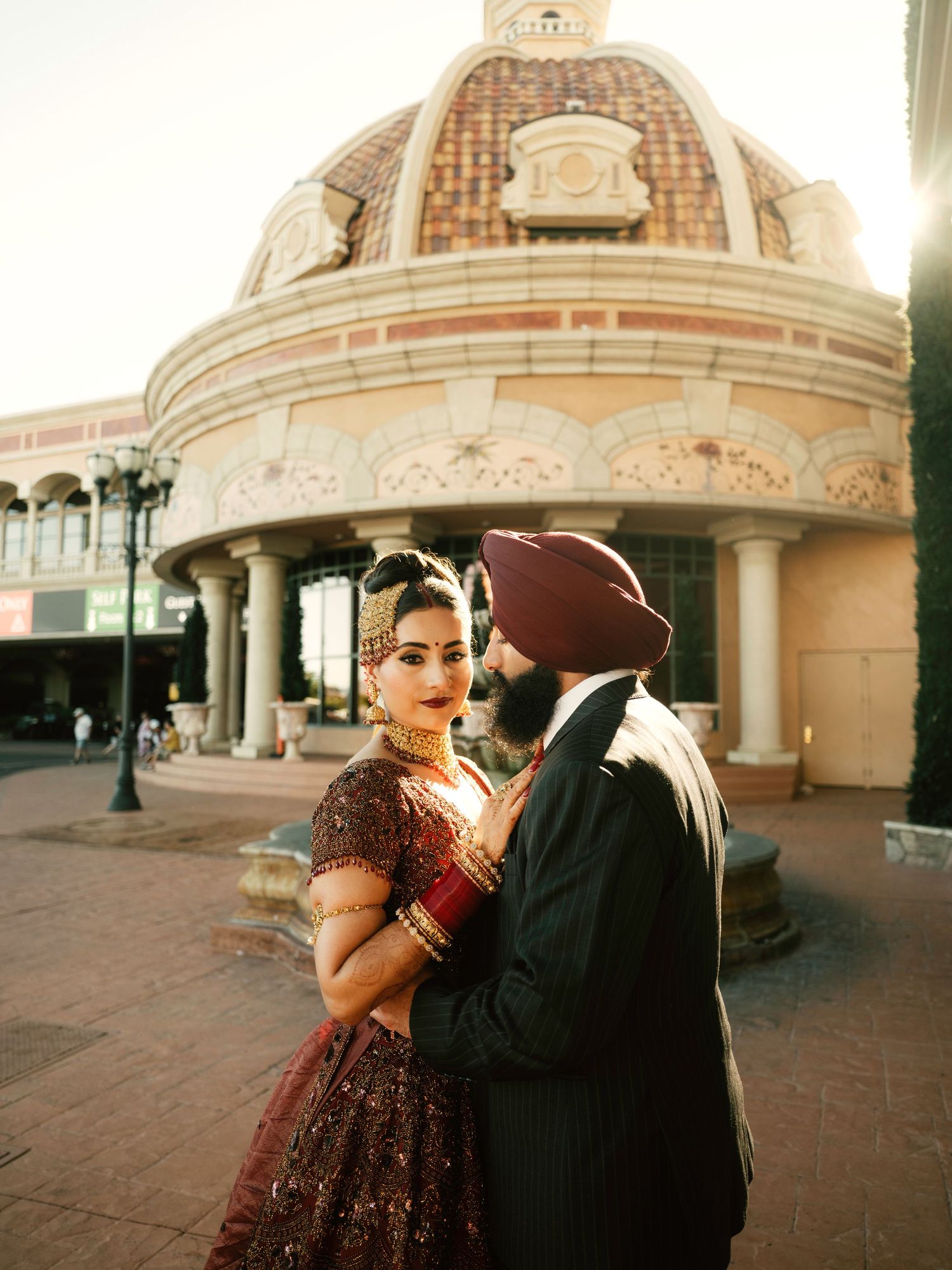 A couple in traditional attire poses romantically outside an ornate domed building with colonial architecture.