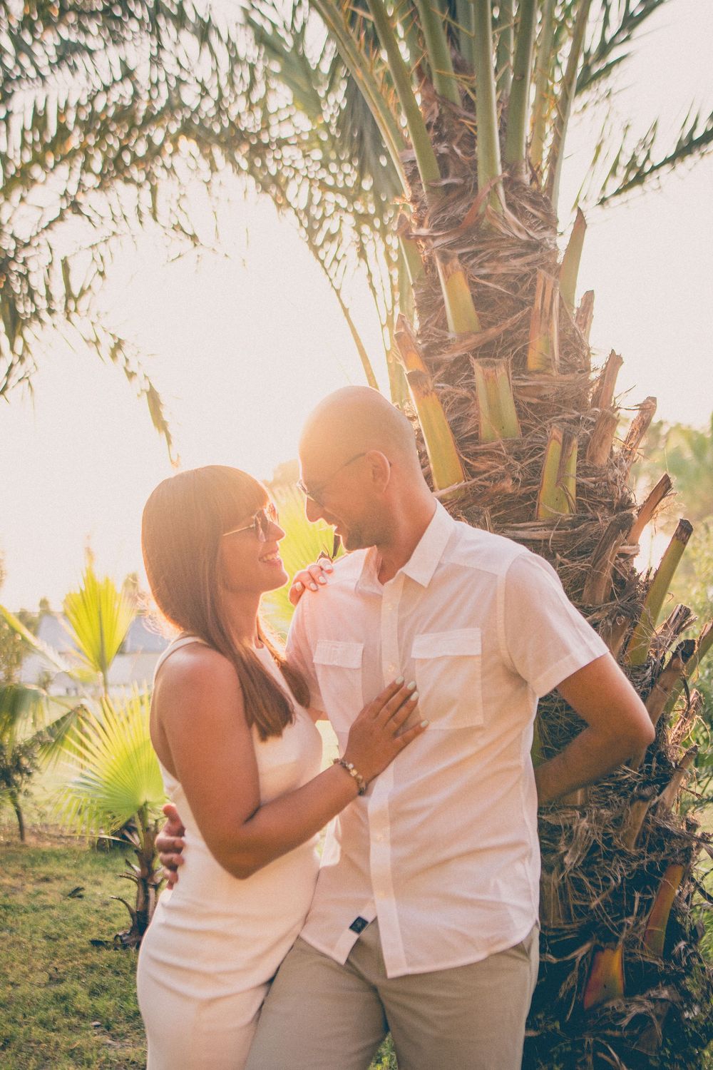 A romantic couple in white outfits share tender moments against a tropical sunset backdrop with palm trees.