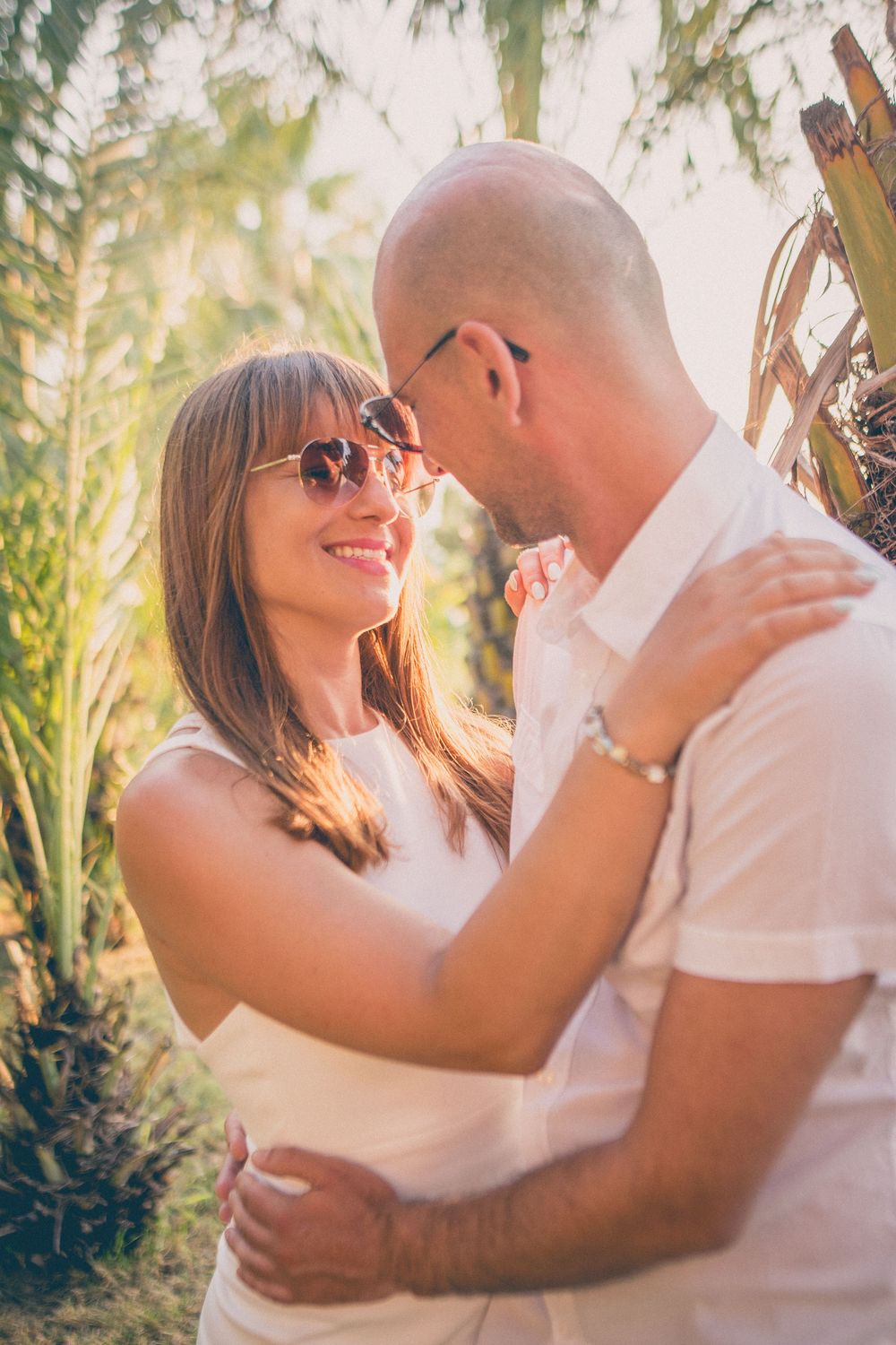 A romantic couple in white outfits share tender moments in a sun-dappled tropical garden setting.