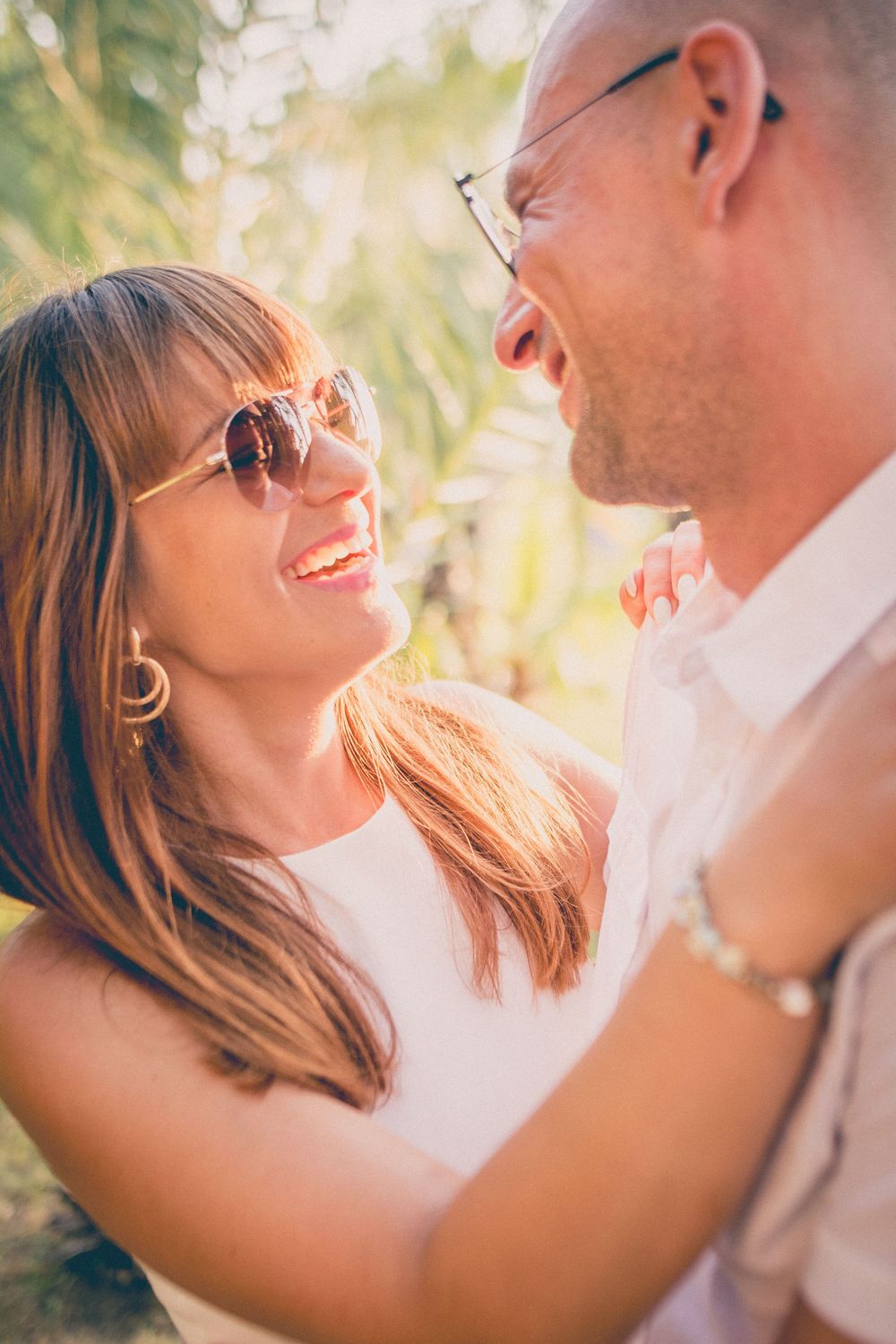 A romantic couple wearing sunglasses smile at each other in warm sunlight during an intimate outdoor moment.