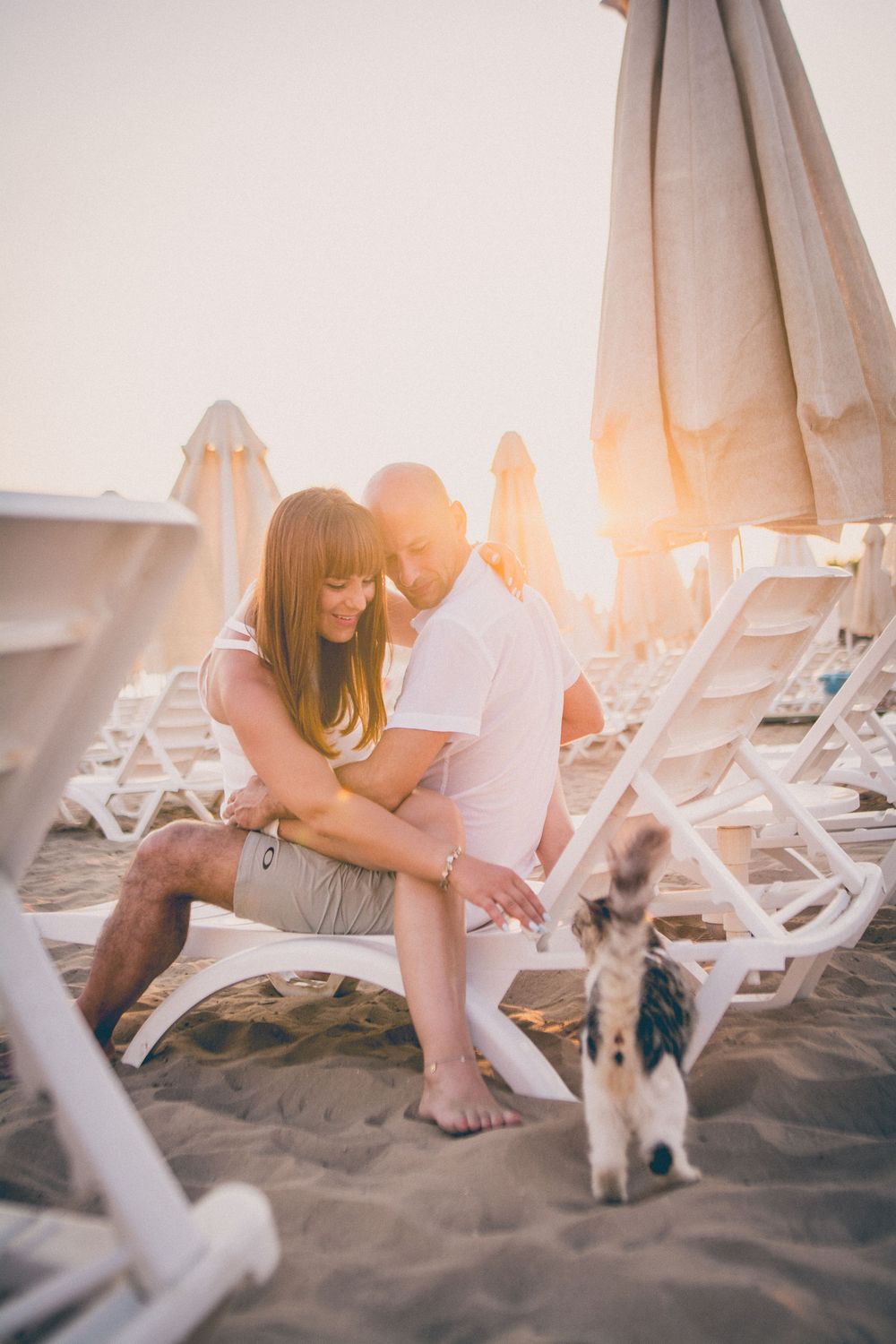 A couple sits on a sandy beach at sunset with beach chairs and umbrellas in the background.