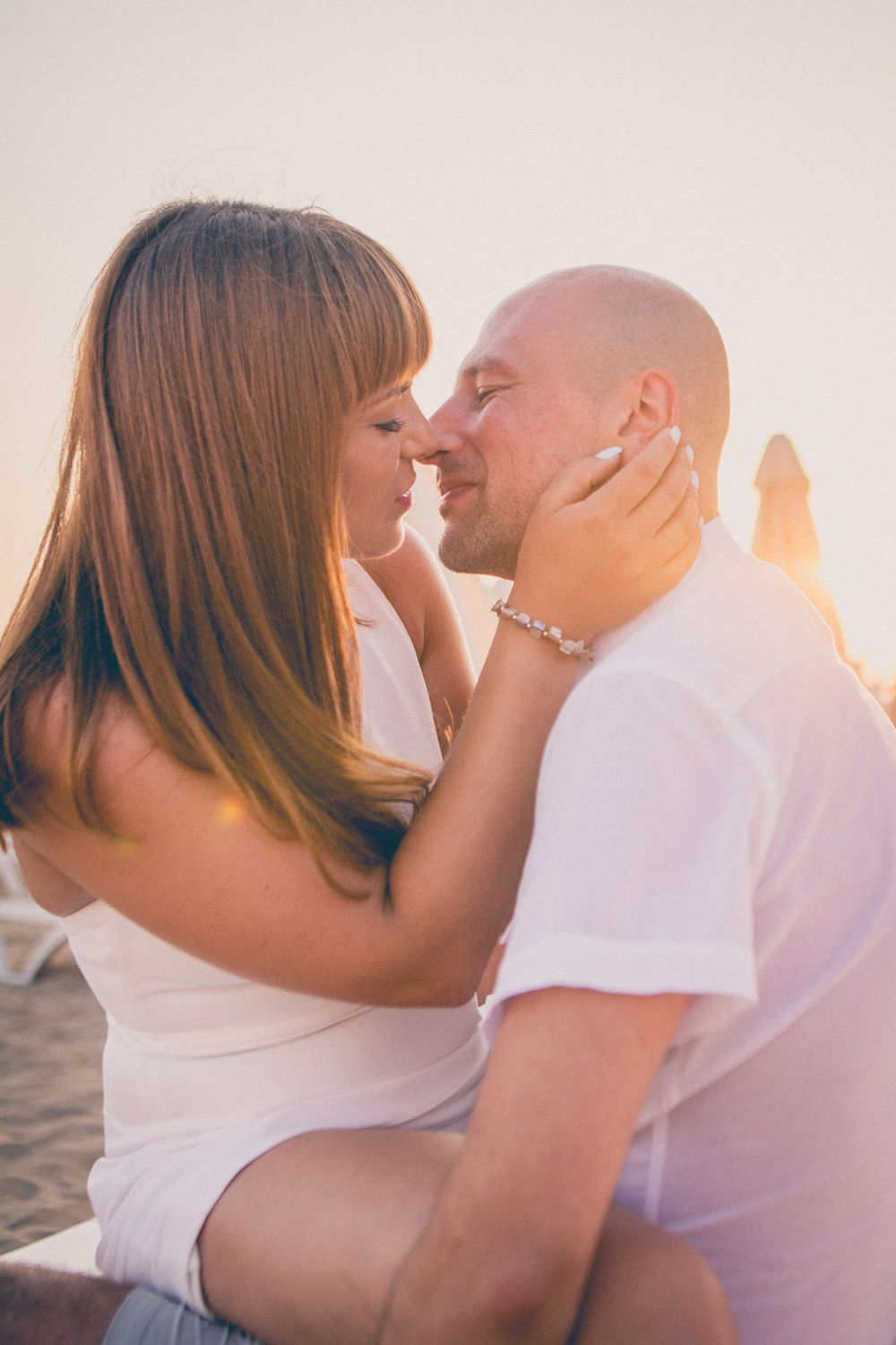 A romantic couple shares an intimate moment at sunset while wearing white outfits on the beach.