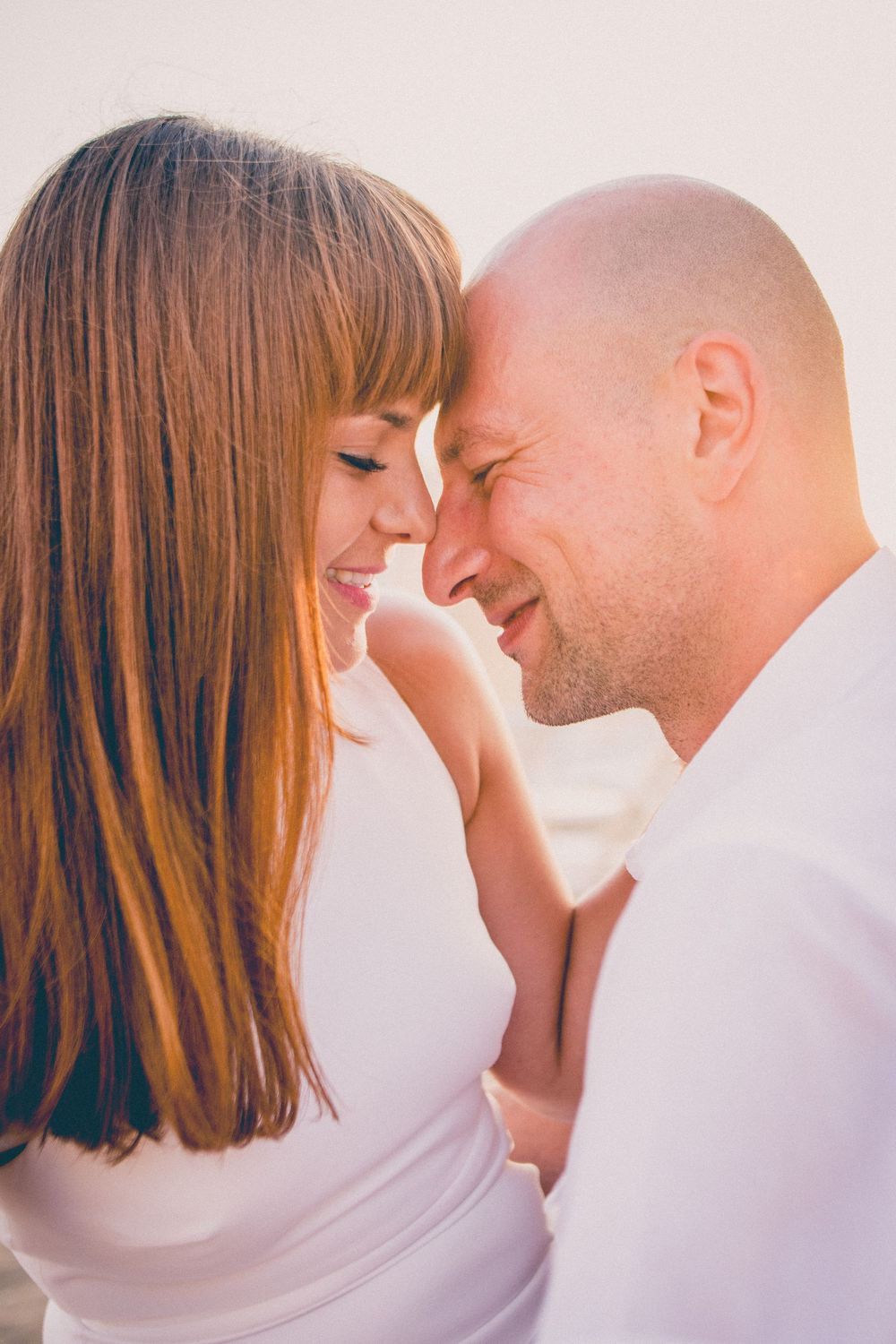 A romantic couple shares an intimate moment with foreheads touching against a bright sunlit background.
