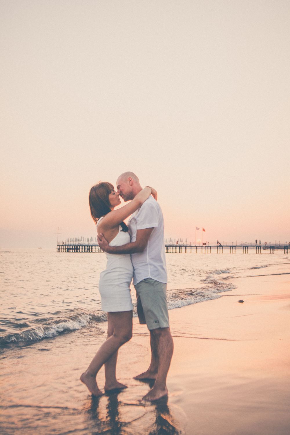 Couple embracing on beach at sunset with pier in background.