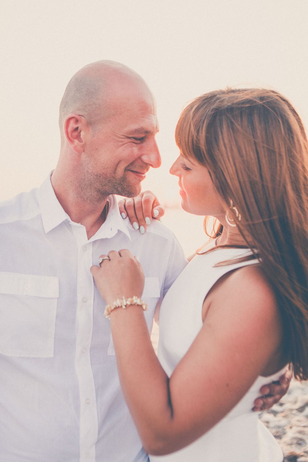 A romantic couple shares tender moments in white attire against a bright sunlit backdrop during a beach photoshoot.