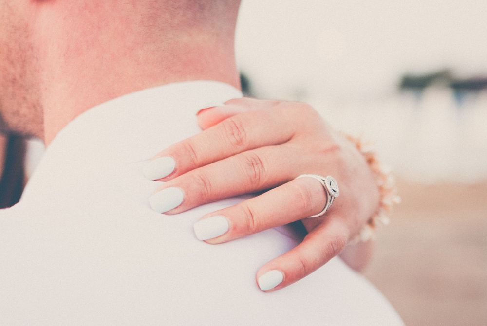 Close-up view of hands with French manicured nails and an engagement ring against a soft blurred background.