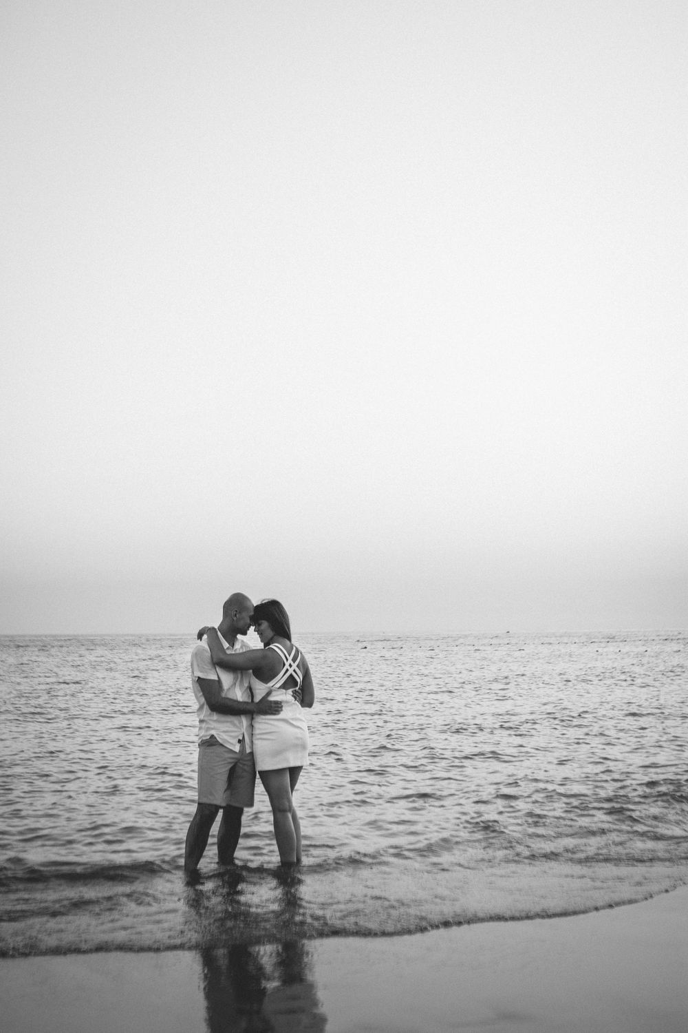 A romantic couple embraces while standing in shallow ocean waves at the beach in black and white photography.