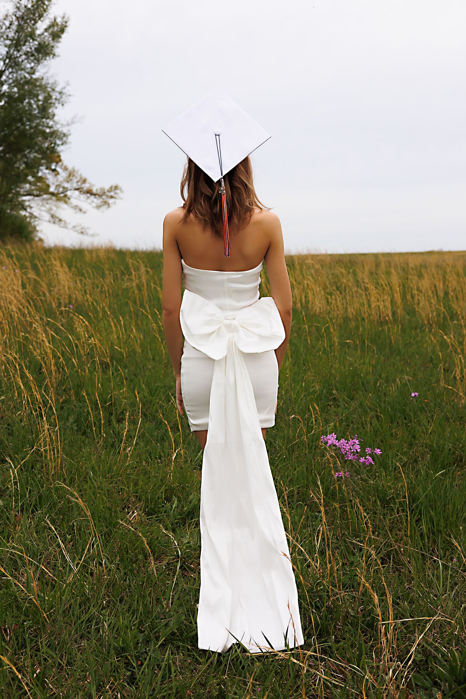 A person in a white dress with a large bow stands in a field of tall grass and wildflowers at sunset.