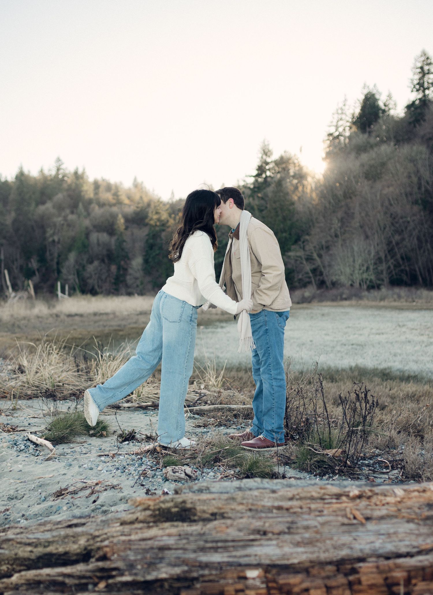 A couple stands together in a grassy field during golden hour while looking down at something on the ground.