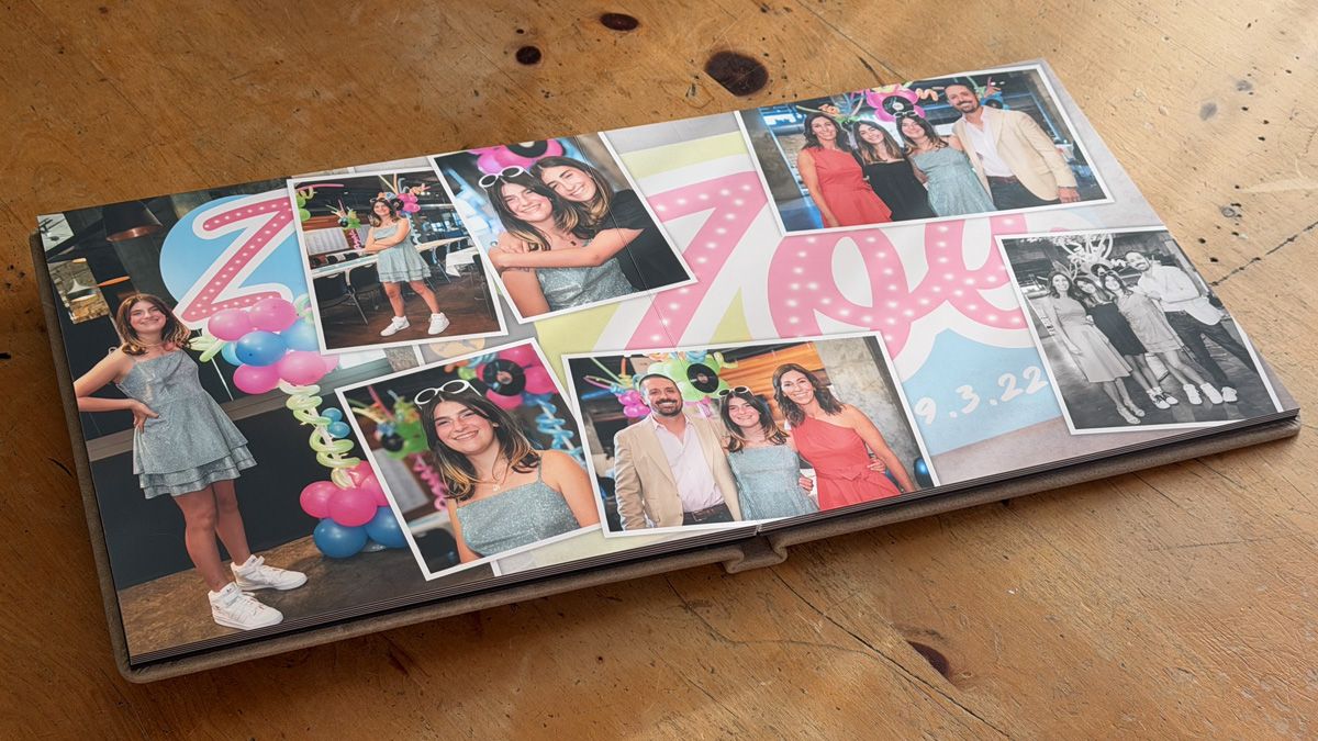 A photo album lies open on a wooden surface showing various personal photographs arranged with pink decorative elements.