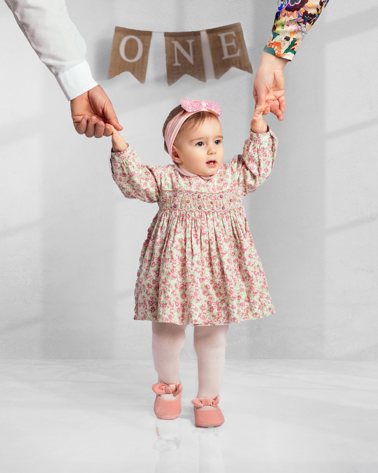Baby girl in floral dress holding parents hands under ONE birthday banner in studio.