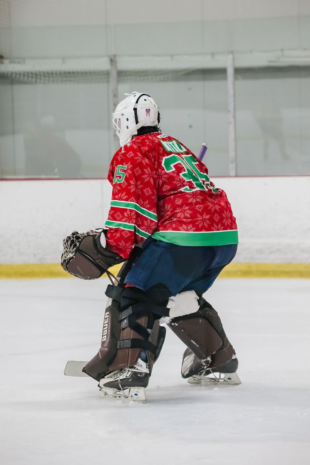 Goalie in red and green uniform crouches in defensive position on ice rink.