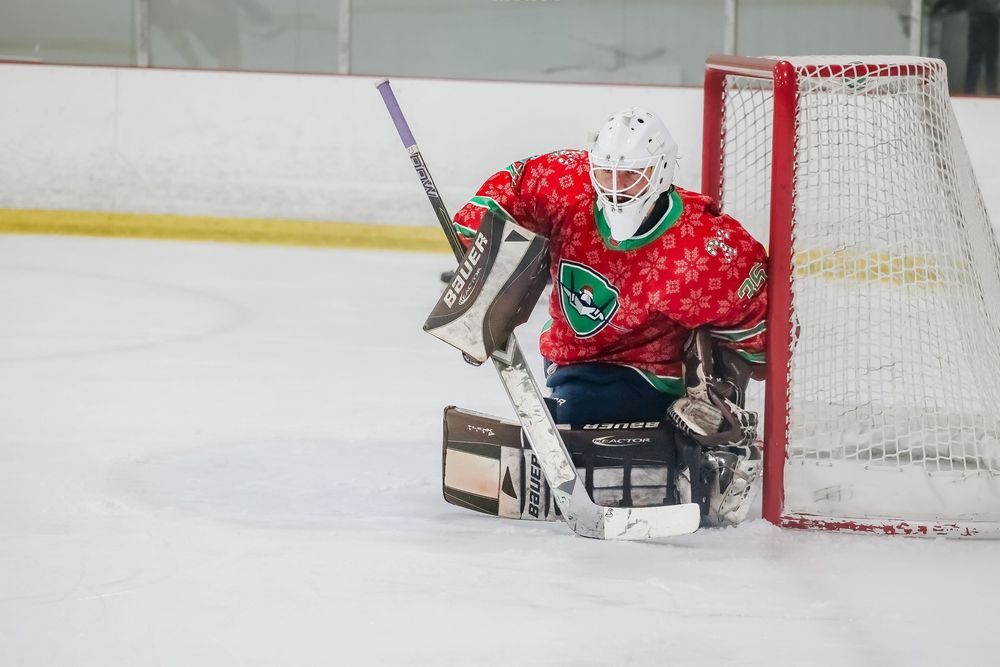 Goalie in red and green uniform defends goal during ice hockey match.