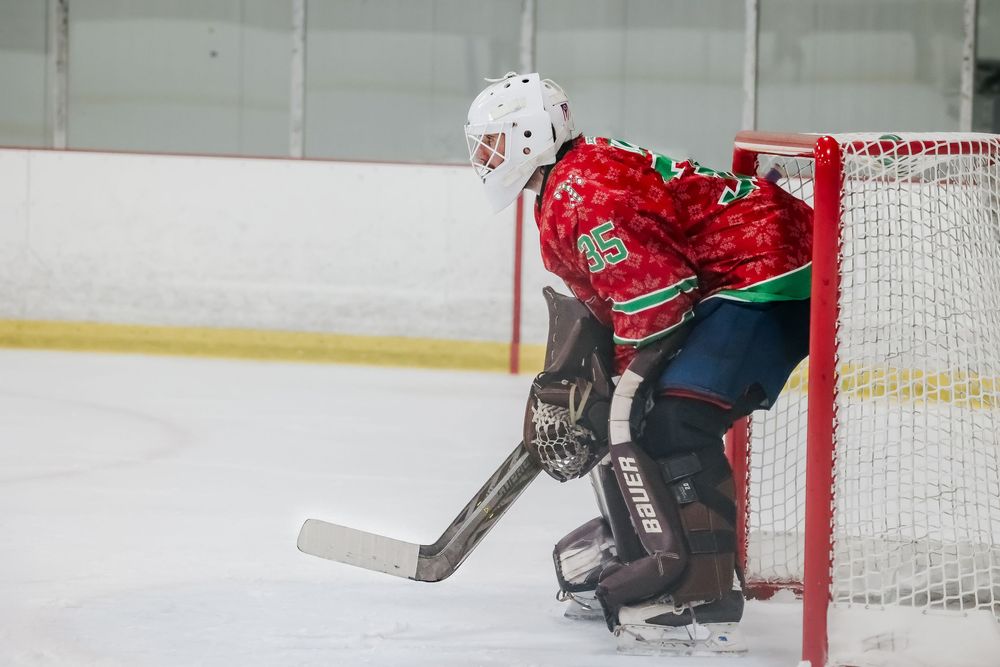 Ice hockey goalie in red and green uniform guards the net during game play.