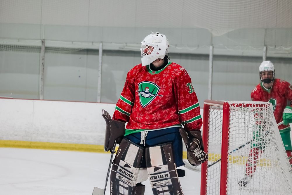 Hockey goalie in red and green uniform crouches in defensive position near goal post.