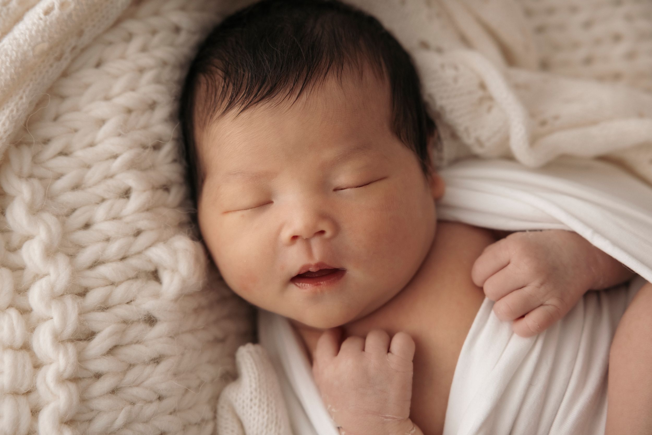 Sleeping newborn baby wrapped in soft white knit blanket during professional photography session.