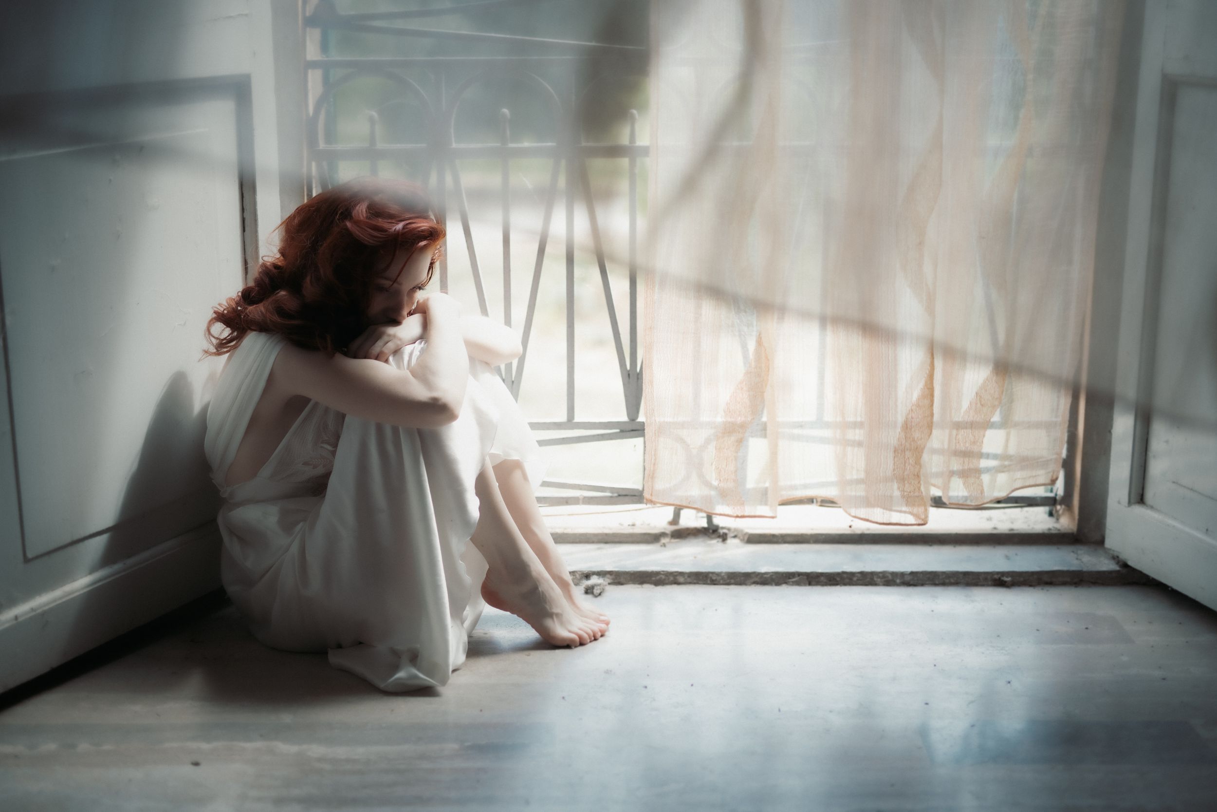 A figure in a flowing white dress sits by a bright window in a minimalist interior space.