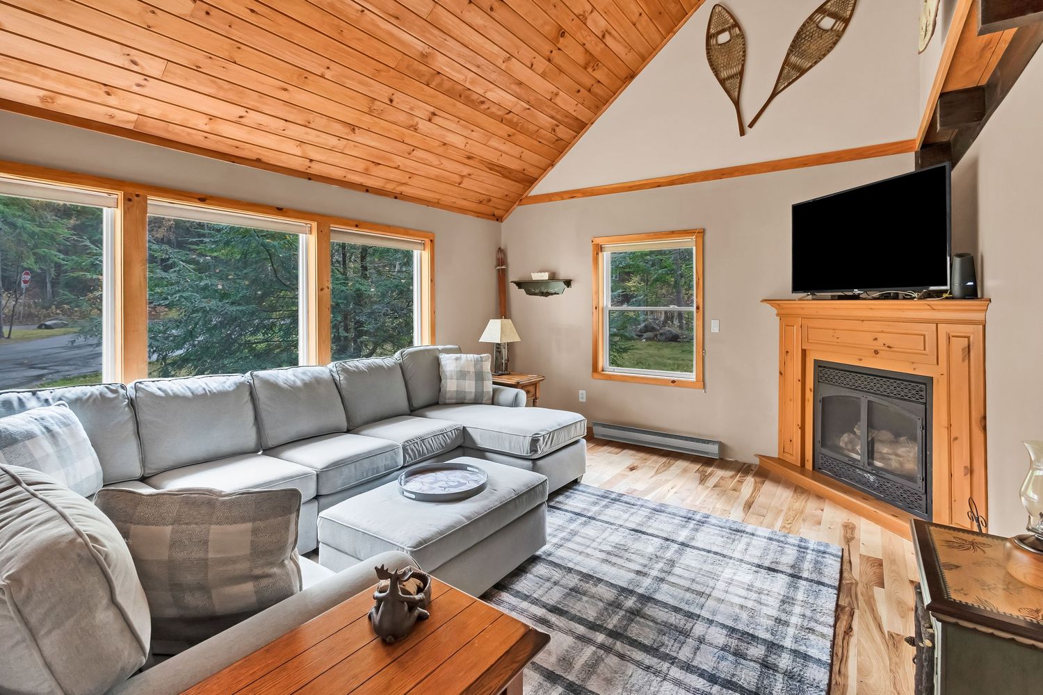 Cozy cabin bedroom with wood paneled ceiling, grey bedding, and natural light streaming through windows.