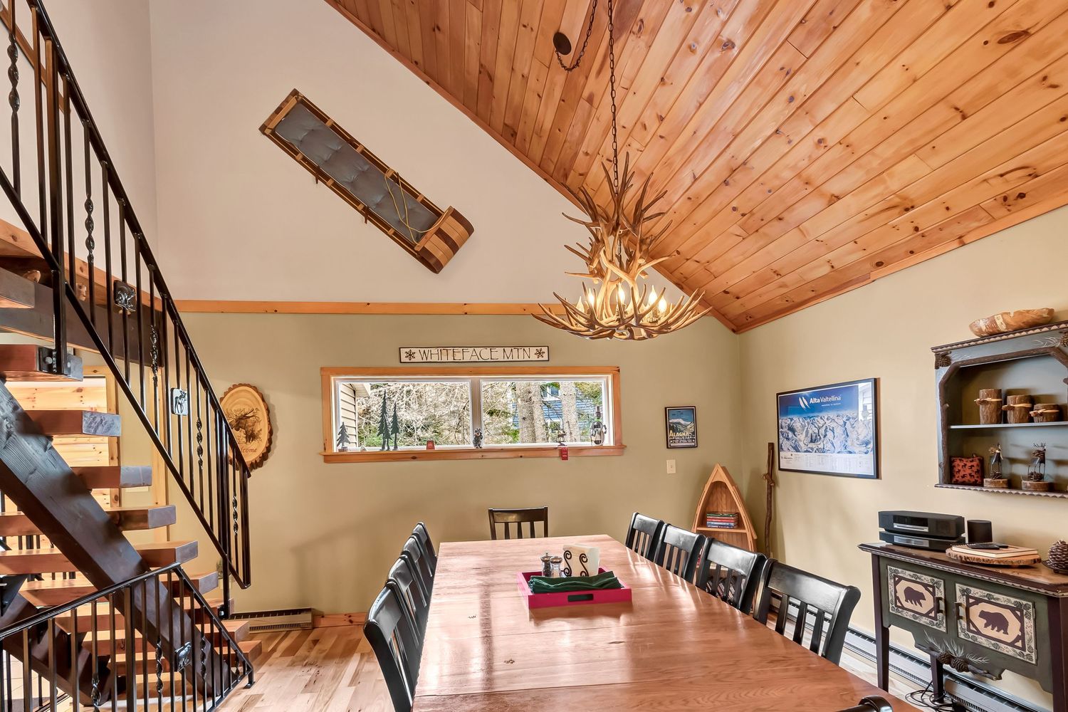 Rustic dining room with wood-paneled vaulted ceiling, long table, and wrought iron staircase in a mountain cabin.