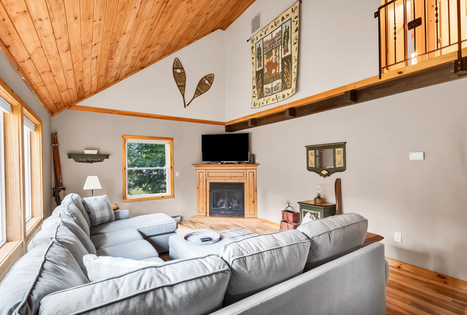 Cozy living room with vaulted wood ceiling, gray sectional sofa, mounted TV, and rustic decor including snowshoes on wall.