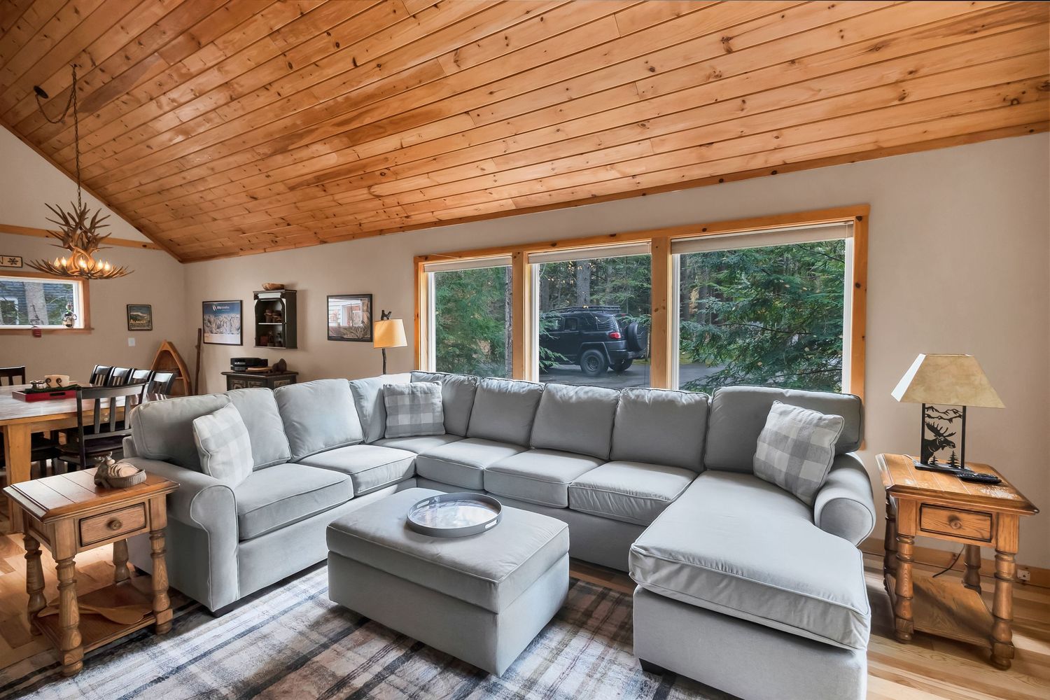 Modern cabin interior with wood paneled ceiling, grey sectional sofa, and large windows overlooking forest views.