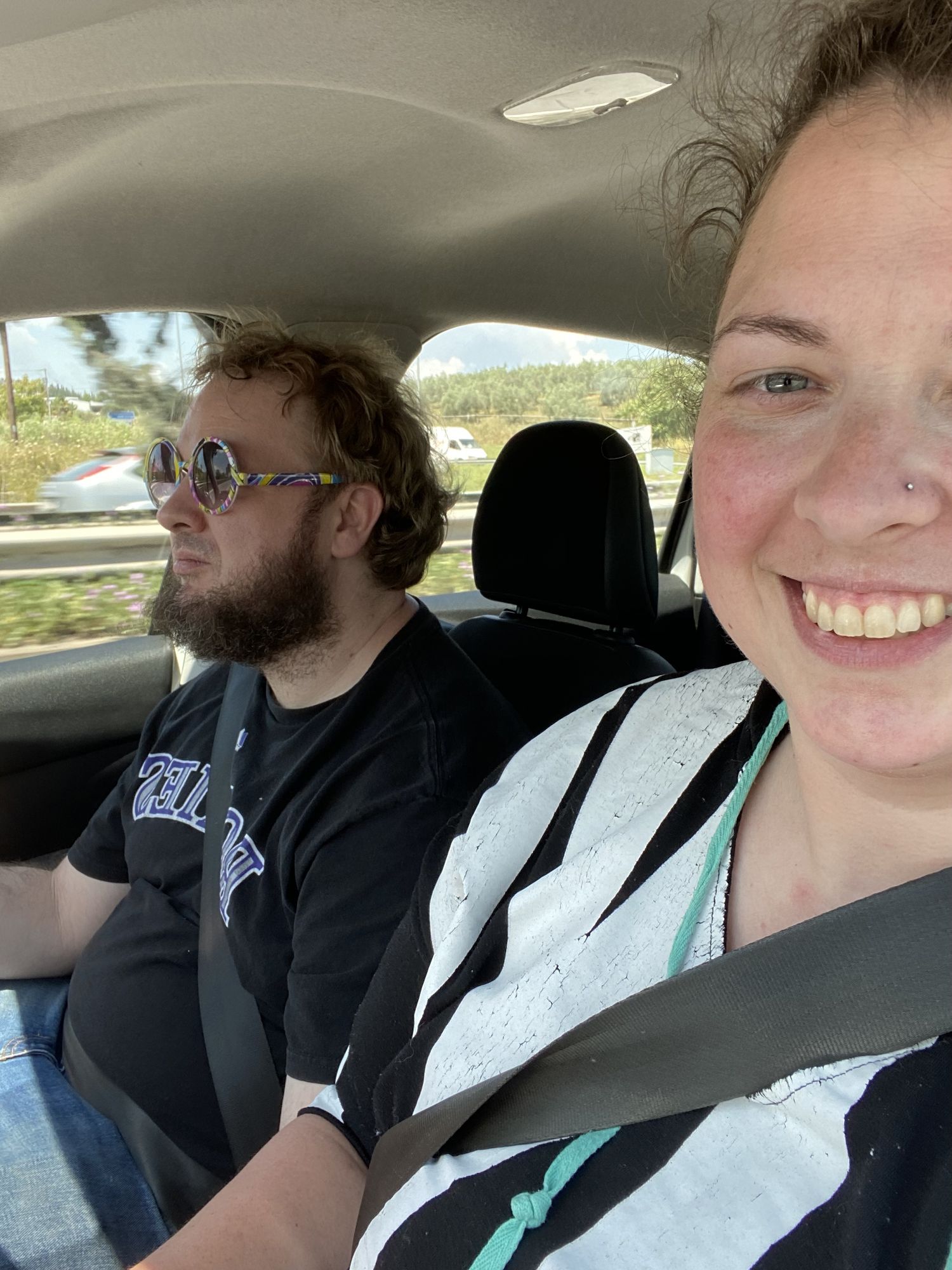 Two people taking a selfie from inside a car on a sunny day while driving down the road.