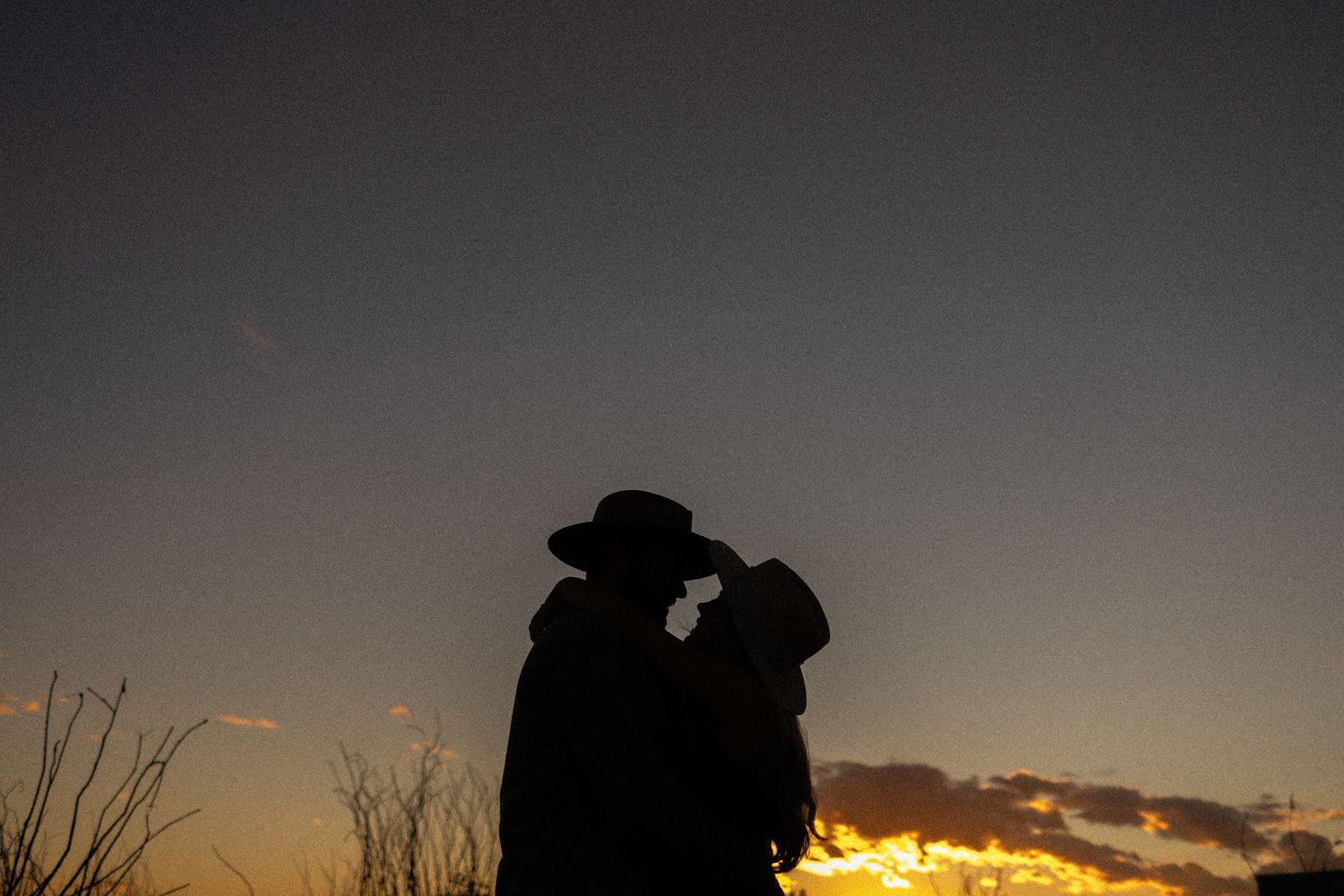 Silhouetted couple in cowboy hats embrace against a dramatic sunset sky with golden clouds.
