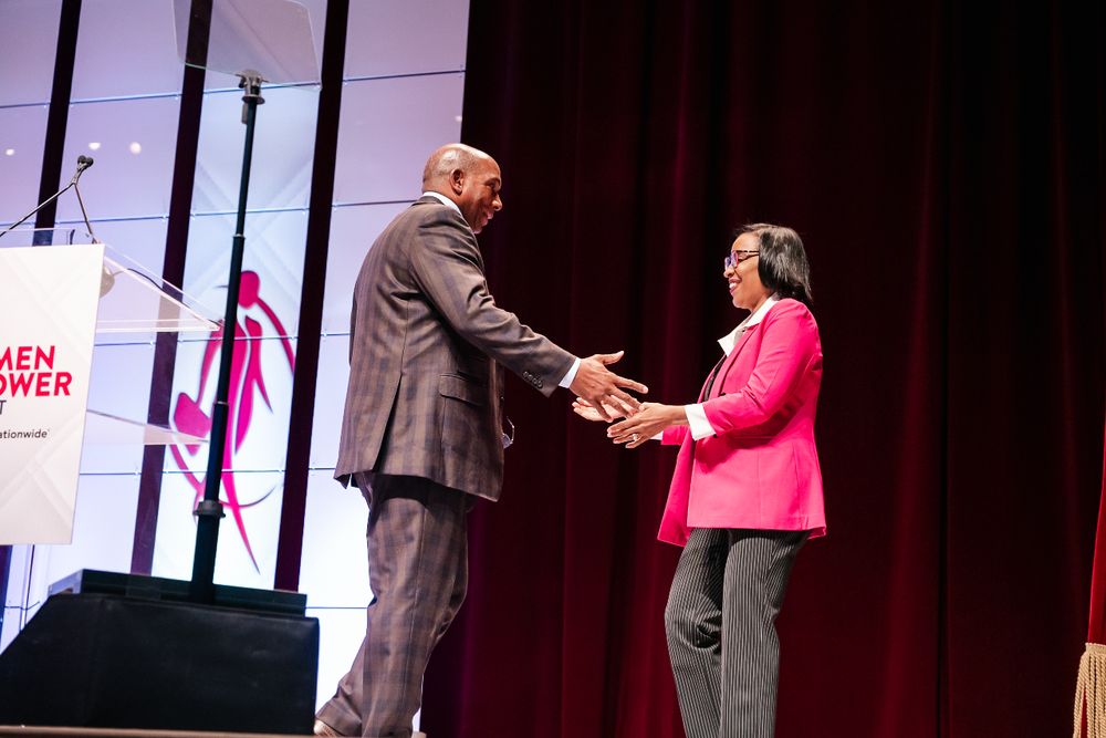 Two people shake hands on stage during an awards ceremony with a red curtain backdrop.
