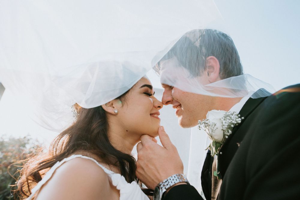 A romantic wedding moment captured under a flowing veil on a sunny day with a soft mountain backdrop.